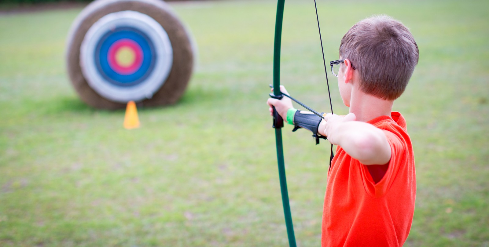 A child wearing glasses and an orange t-shirt draws a bow and arrow while aiming at a circular archery target in a grassy field. The target has traditional blue, red, and yellow rings