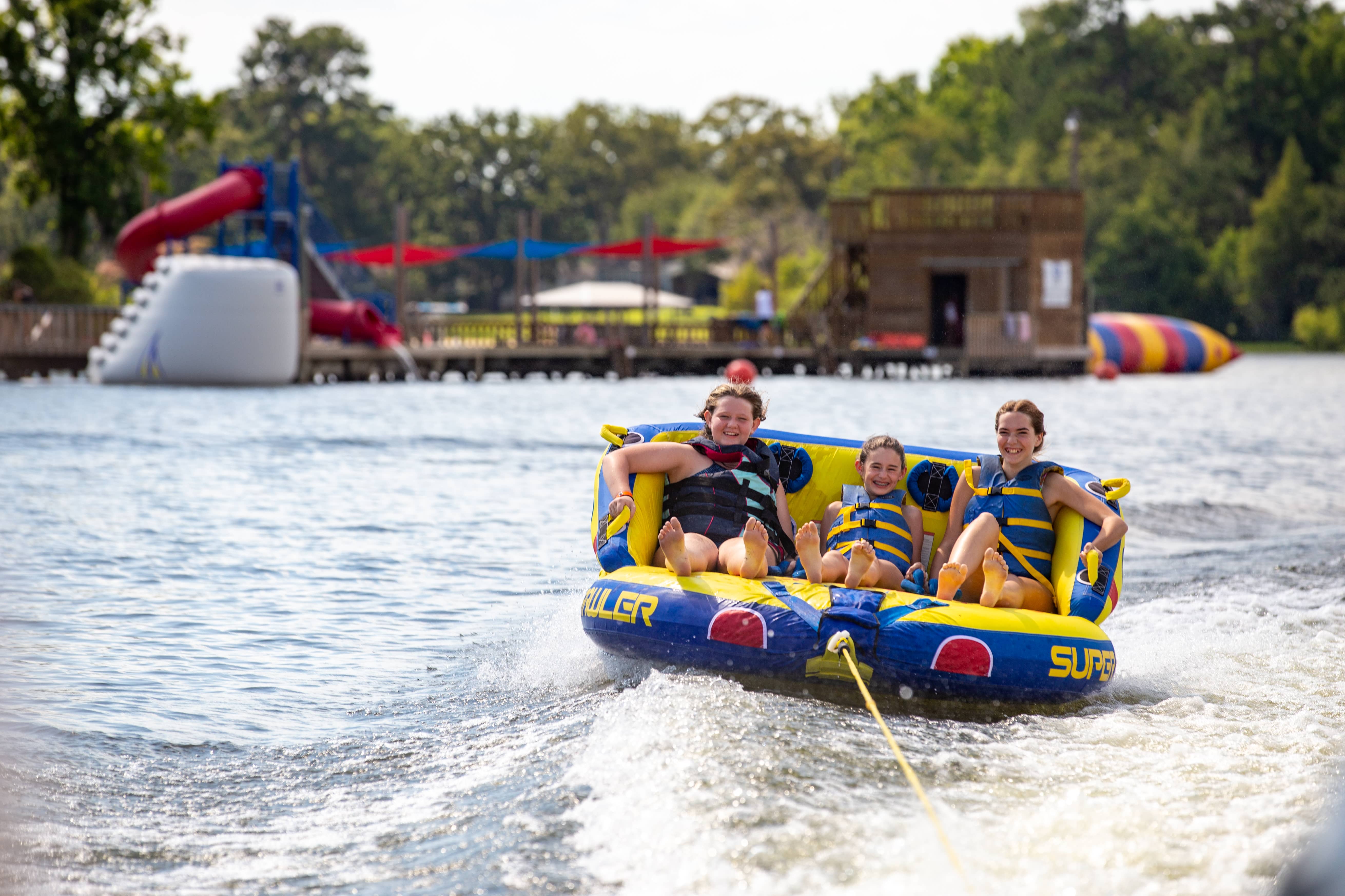 A child wearing glasses and an orange t-shirt draws a bow and arrow while aiming at a circular archery target in a grassy field. The target has traditional blue, red, and yellow rings